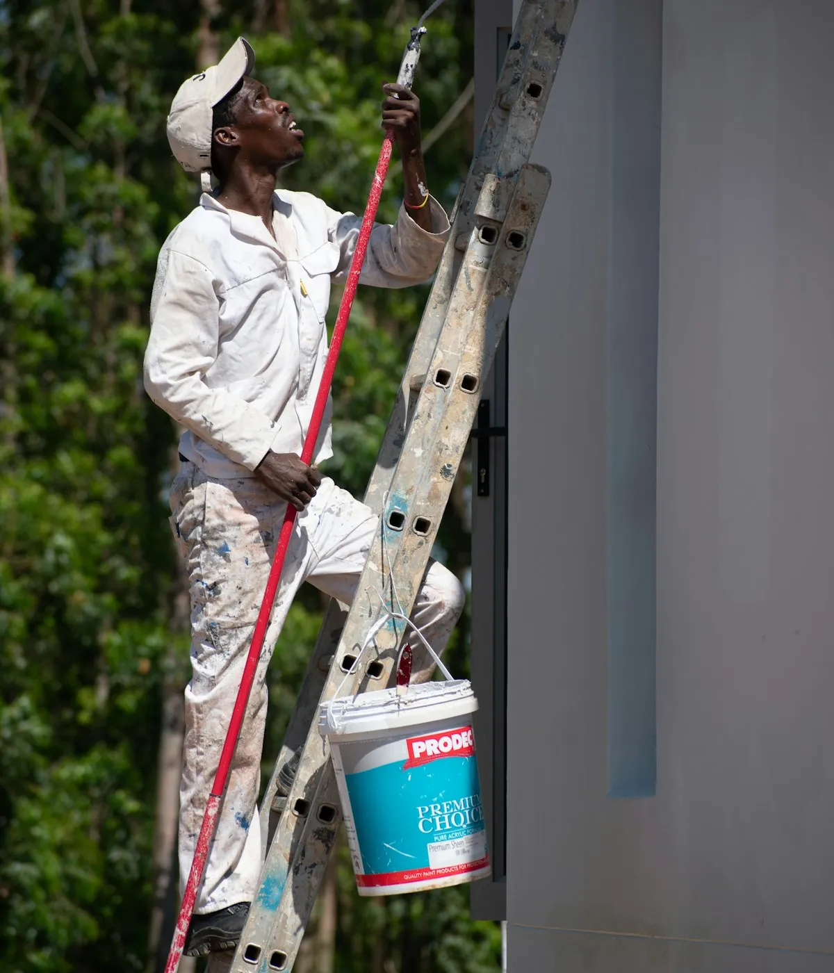 A painter on an extension ladder cutting in beside a window, paint pail hooked to the rung
