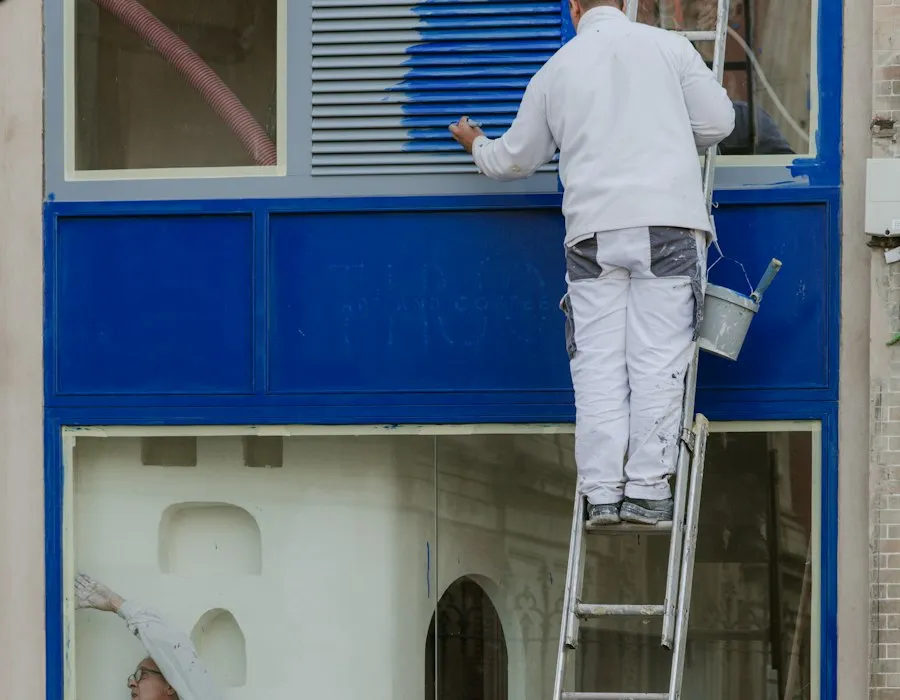 A painter working on the trim around an exterior window from a ladder.