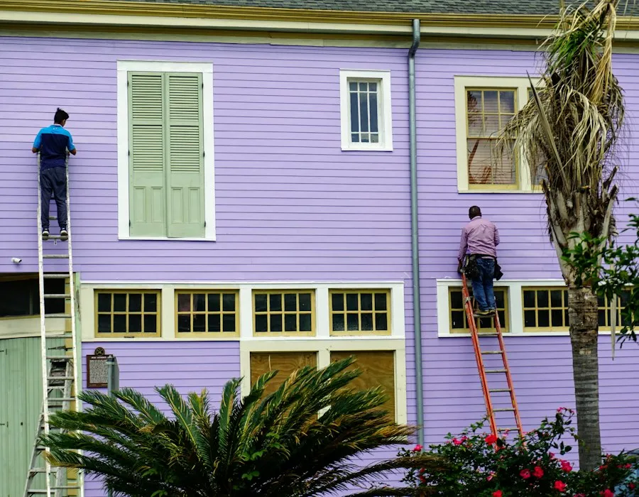 Two painters working from ladders on the upper wall of an older house.